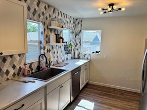A kitchen with a stone backsplash, white cabinets, a stainless steel sink, and a window with a view of a house.