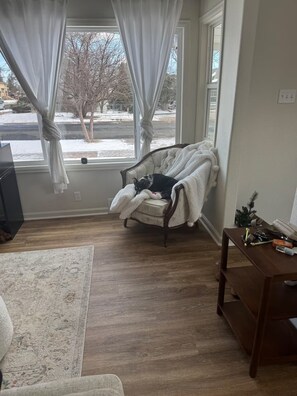 A room with a large window, a chair with a blanket, a wooden floor, and a small table with items. - Golden Hour Getaway, tucked less than a mile from Frontier Park and downtown! (Cheyenne)