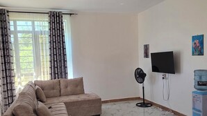 A living room with a sectional sofa, a flat-screen TV, a standing fan, and a water dispenser. - MIHAMBOS RESIDENCE (Msambweni)