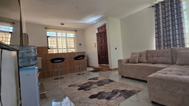 A living room with a sofa, a coffee table, and a patterned rug. There is a water dispenser and a door leading to another room. - MIHAMBOS RESIDENCE (Msambweni)