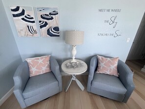 Ein Zimmer mit zwei grauen Sessel, einem kleinen runden Tisch mit Lampe und Wandbildern abstrakter Formen.