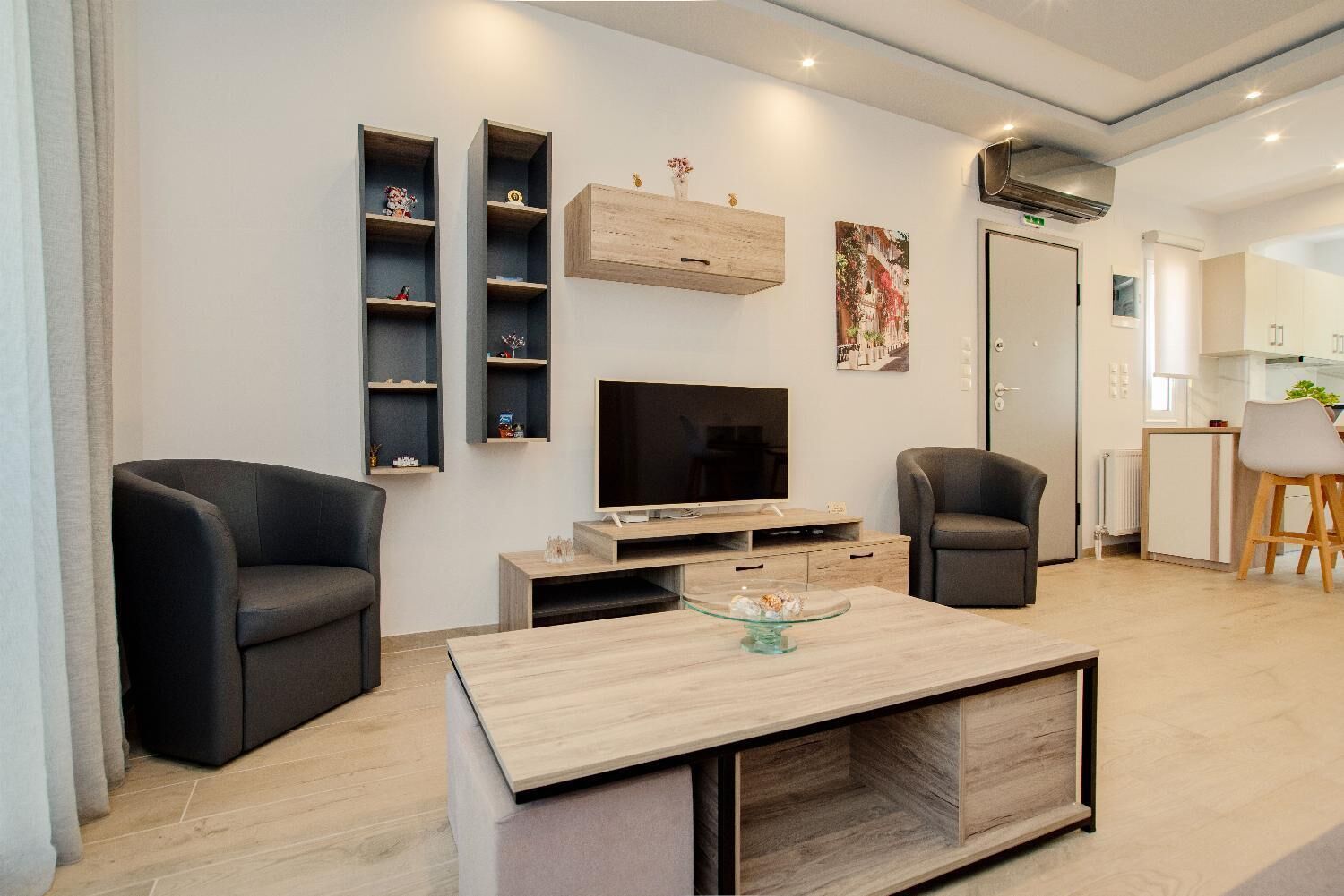 Ein modernes Wohnzimmer mit Flachbildfernseher, zwei schwarzen Sesseln, einem Holztisch und einer Einbauschränke mit Dekorationselementen.