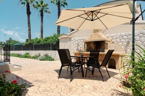 An outdoor patio with a stone-tiled floor, a table with chairs, a barbecue grill, and a large umbrella. - Villa Boltado - Two Bedroom Villa, Sleeps 4 (Lefkimmi)