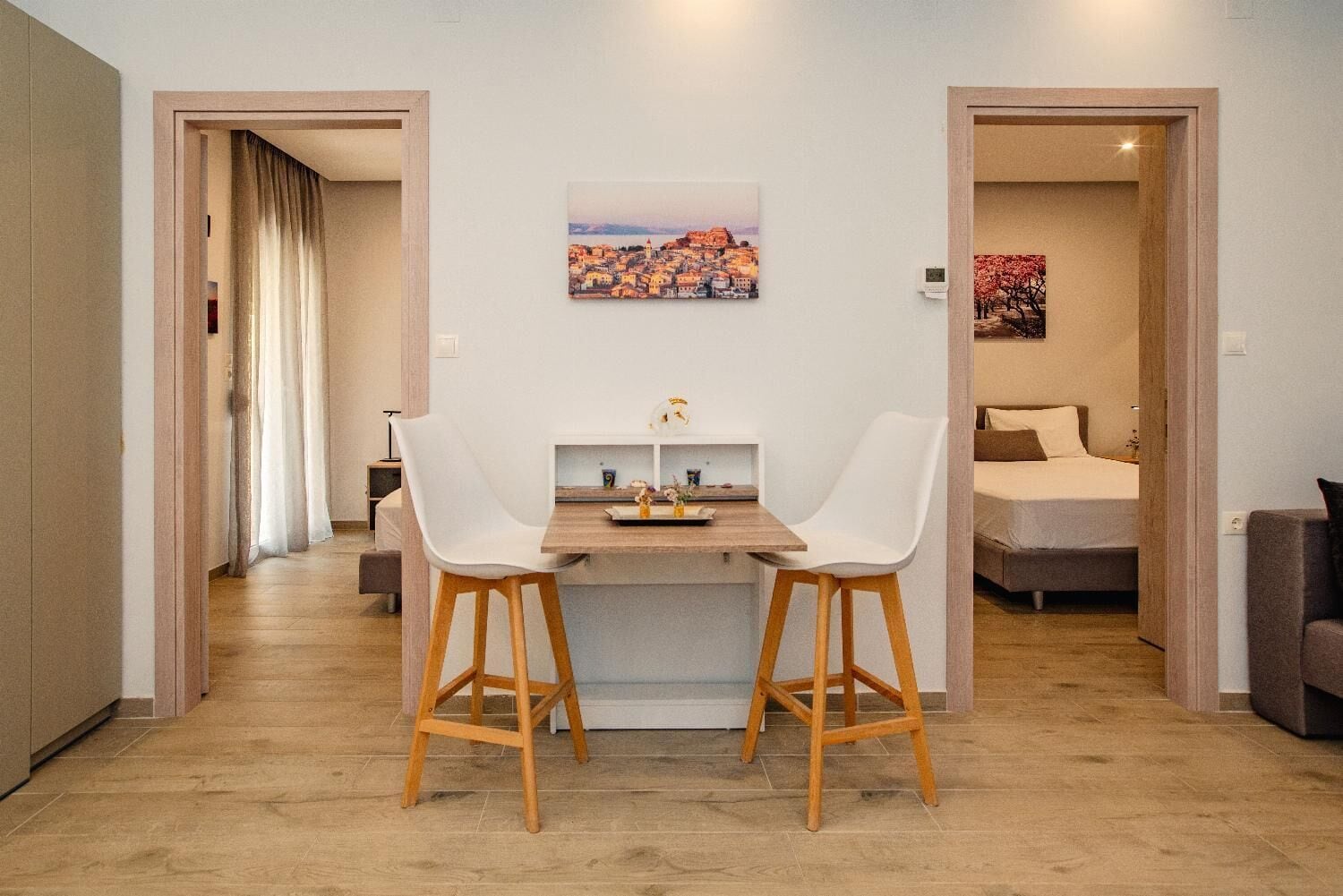 A modern room with a dining area featuring two white chairs and a wooden table, a bed with a brown headboard, and a painting of a cityscape on the wall.