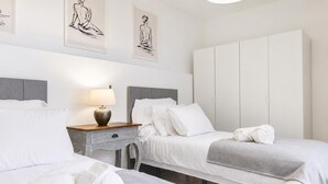 Quarto de hotel com duas camas, uma mesa de cabeceira com abajur, guarda-roupa e uma obra de arte na parede.