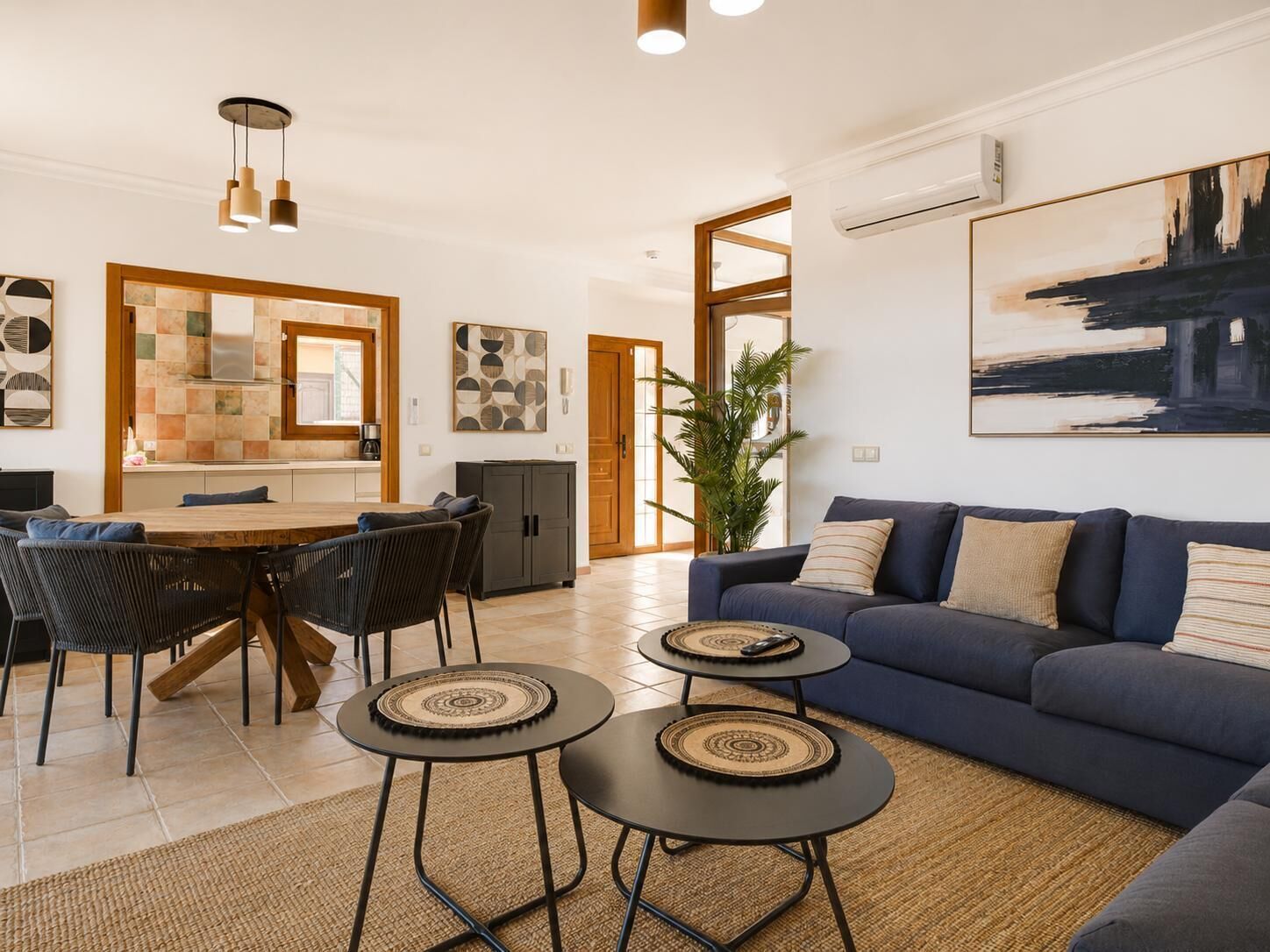Ein modernes Wohnzimmer mit einem blauen Sofa, zwei kleinen runden Tischen, einer Essecke mit Holztisch und Stühlen sowie einer Küche mit gefliestem Spritzschutz.