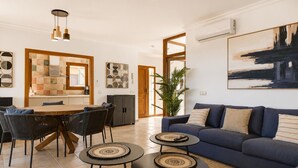 Uma sala de estar moderna com um sofá azul, duas mesas redondas pequenas, uma área de jantar com uma mesa e cadeiras de madeira, e uma cozinha com painel de azulejos.