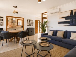 Ein modernes Wohnzimmer mit einem blauen Sofa, zwei kleinen runden Tischen, einer Essecke mit Holztisch und Stühlen sowie einer Küche mit gefliestem Spritzschutz.