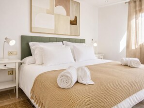 Un lit bien fait, avec du linge de lit blanc et un plaid beige, deux lampes de chevet et un tableau abstrait accroché au mur.