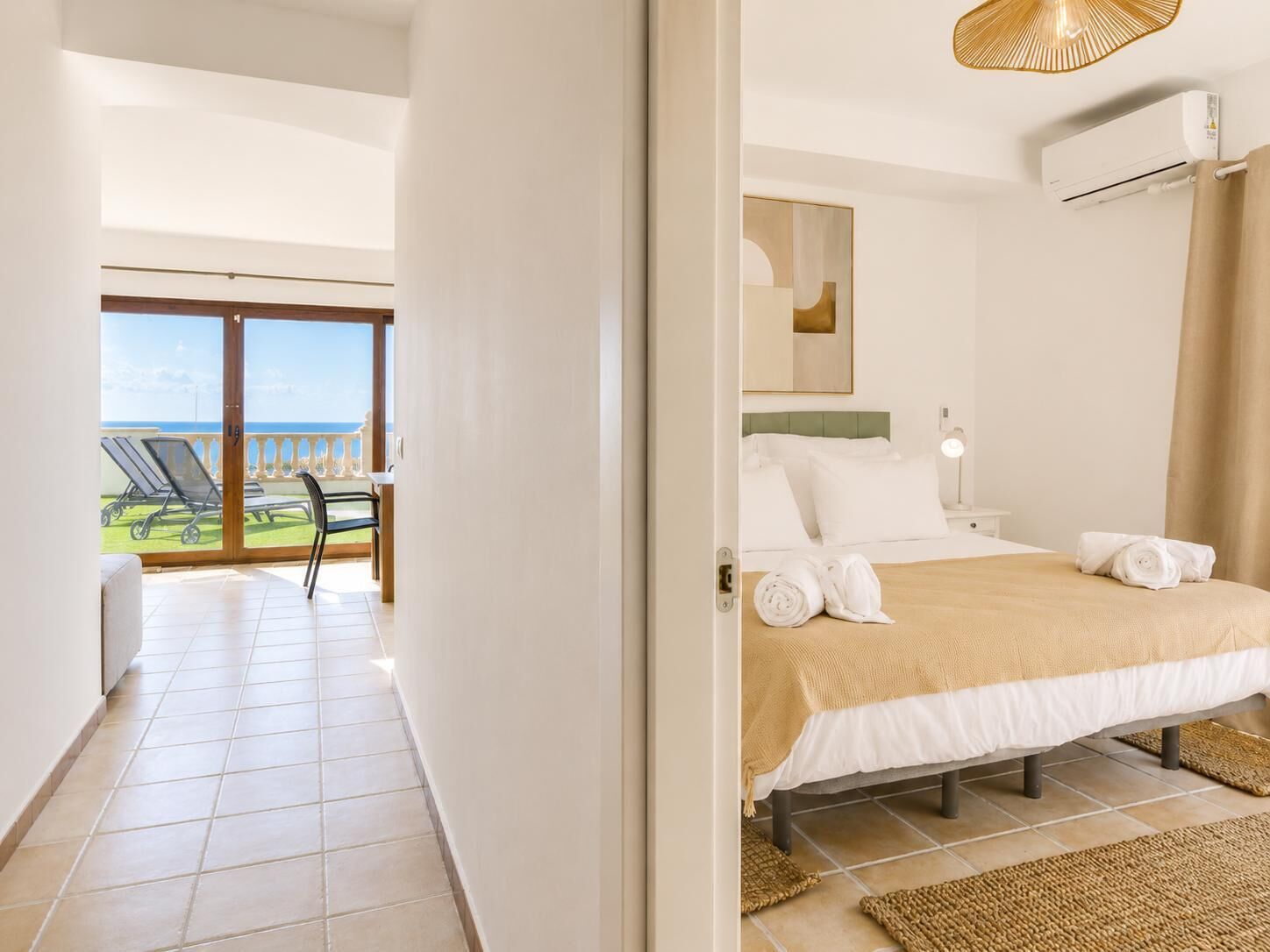 A hotel room with a bed, a view of the sea, a balcony with chairs, and an air conditioning unit.
