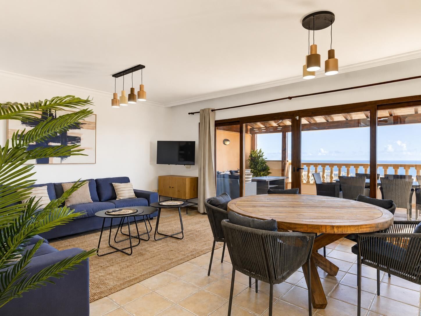 A modern living room with a sofa, a round wooden table, and chairs, a television, and a balcony with outdoor seating.