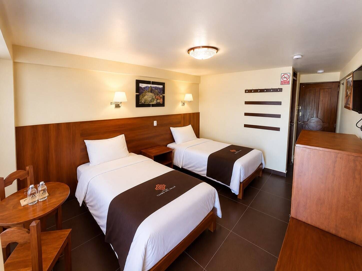 A hotel room with two beds, a wooden headboard, a small table with a chair, a wall-mounted TV, and a door with a sign.