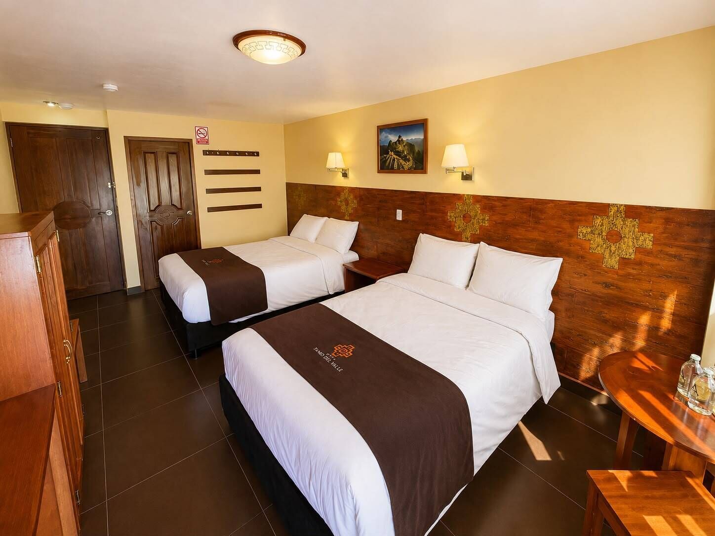 Habitación de hotel con dos camas, cabecera de madera, mesita de noche, escritorio y lámpara de pared.
