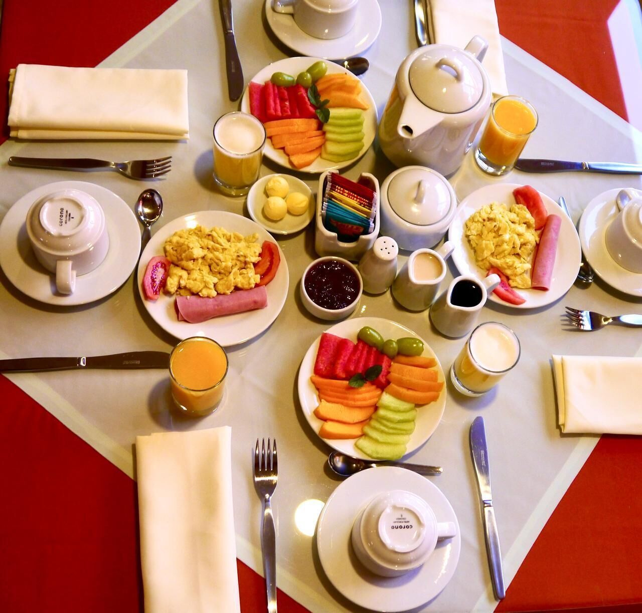 A breakfast setting with plates of eggs, ham, and fruit, a teapot, cups, and a variety of condiments.
