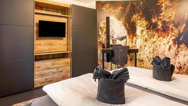 Una camera d'albergo moderna con un letto grande, una TV a schermo piatto montata su un pannello di legno, una scrivania con una sedia e un muro decorato con un murales strutturato.
