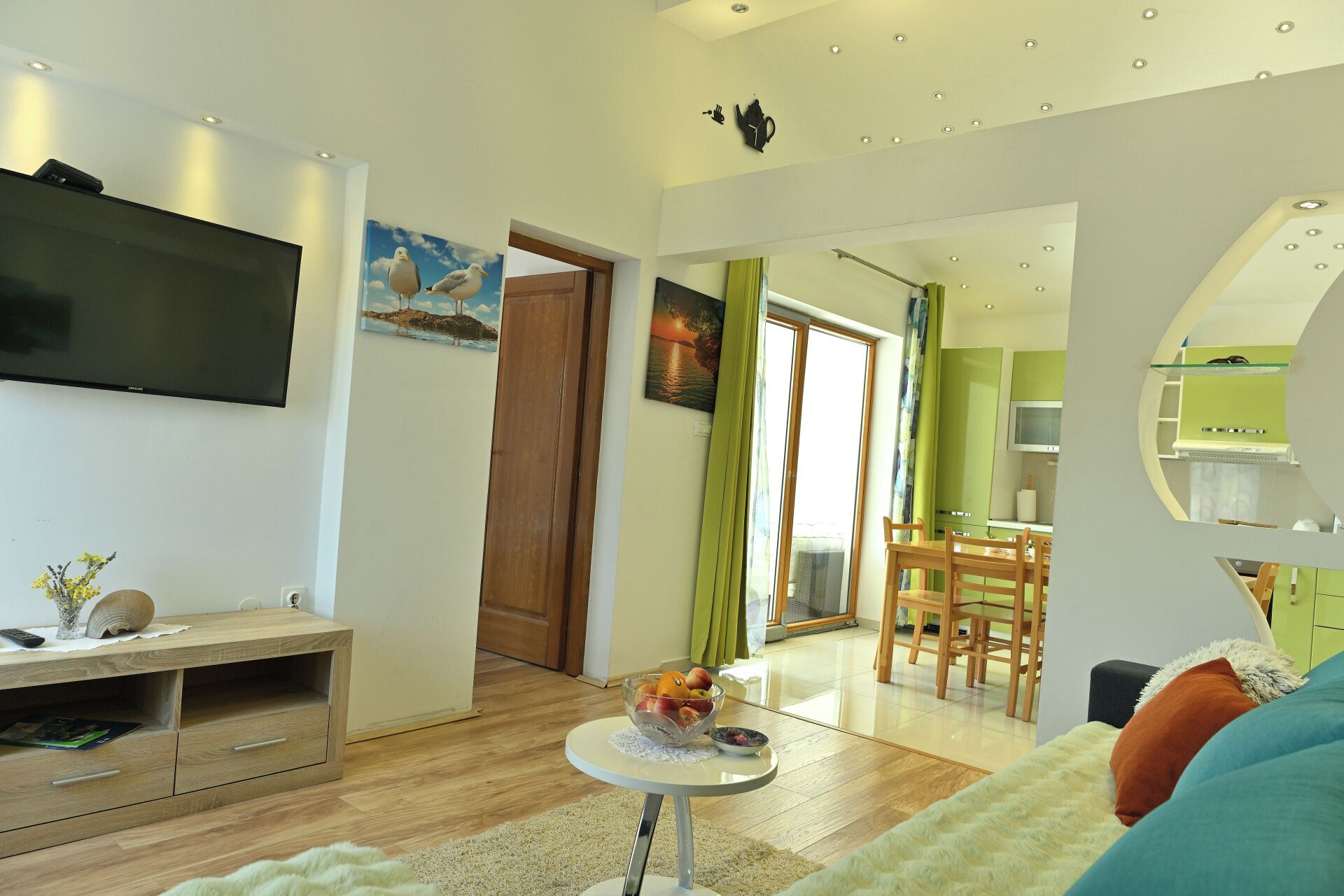 Een moderne woonkamer met een flatscreen televisie, houten meubels, een eethoek met een groene tafel, en een keuken met een magnetron.