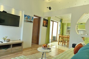 Een moderne woonkamer met een flatscreen televisie, houten meubels, een eethoek met een groene tafel, en een keuken met een magnetron.