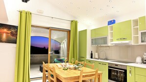 Een moderne keuken met groene kastjes, een eethoek met een houten tafel en stoelen, en uitzicht op de zonsondergang door een schuifdeur.