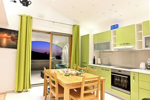 Eine moderne Küche mit grünen Schränken, ein Essbereich mit einem Holztisch und Stühlen sowie Blick auf den Sonnenuntergang durch eine Glastür.