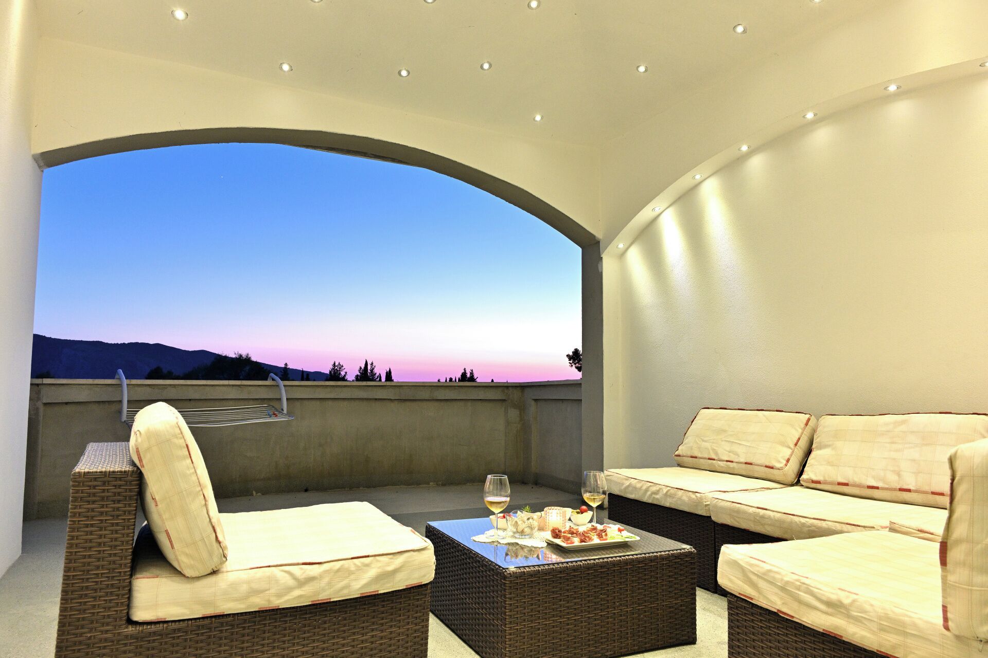 Un espace extérieur aménagé avec des meubles en osier et des coussins, une table avec des verres et de la nourriture, et une vue sur le ciel et le paysage.