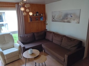 Ein Wohnzimmer mit einer Couch, einem Sessel, zwei kleinen Tischen, einem Bücherregal und einem Bild an der Wand.