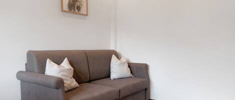 A brown sofa with two pillows against a white wall, a framed picture above it, and a tiled floor.