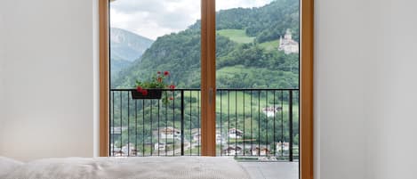 Una habitación con cama, ventana con vistas a un paisaje montañoso y balcón con barandilla y maceta.