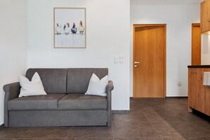 A grey sofa with white cushions in a room with a wooden door and a framed artwork of chickens on the wall. - Apartment 'Trostburg' with Mountain View and Wi-Fi (Barbian)