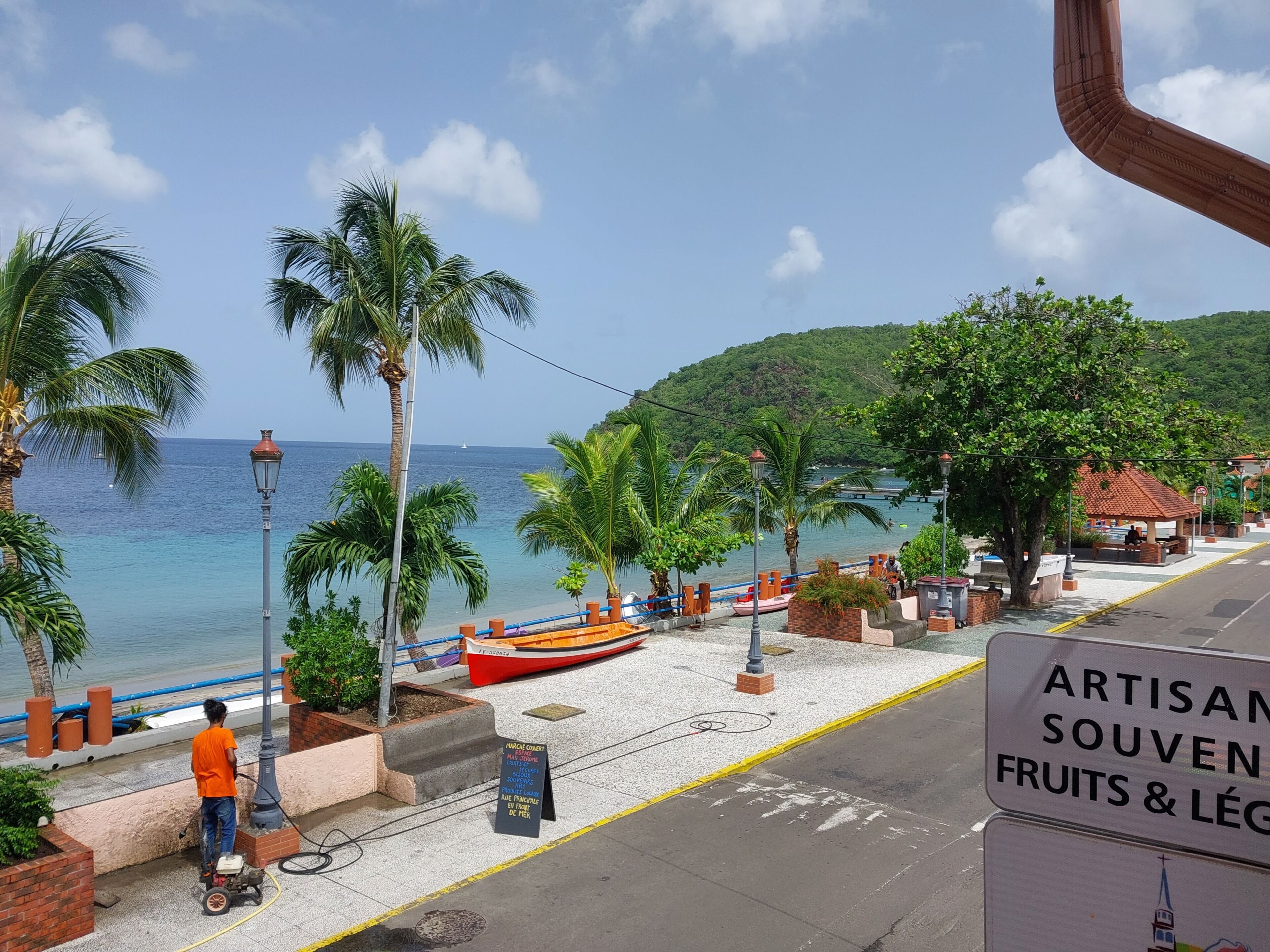 A coastal street with a view of the sea, a red boat, and a sign for 'ARTISAN SOUVENIR FRUITS & LÉGUMES.'