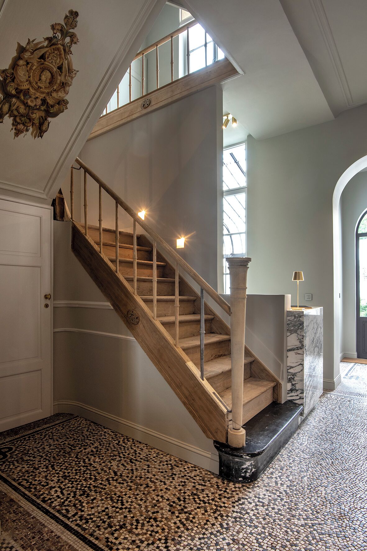 A grand staircase with wooden steps and a marble landing, leading to a room with a large window and a decorative wall piece.
