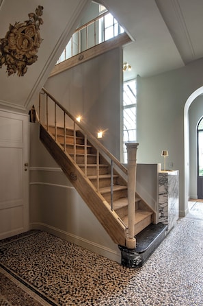 Una imponente escalera de madera con un descansillo de mármol que conduce a un espacio con un amplio ventanal y un elemento decorativo en la pared.