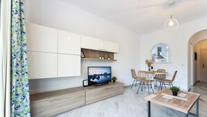 Een moderne woonkamer met een flatscreen televisie, houten kasten, een eethoek met rieten stoelen en een hanglamp.