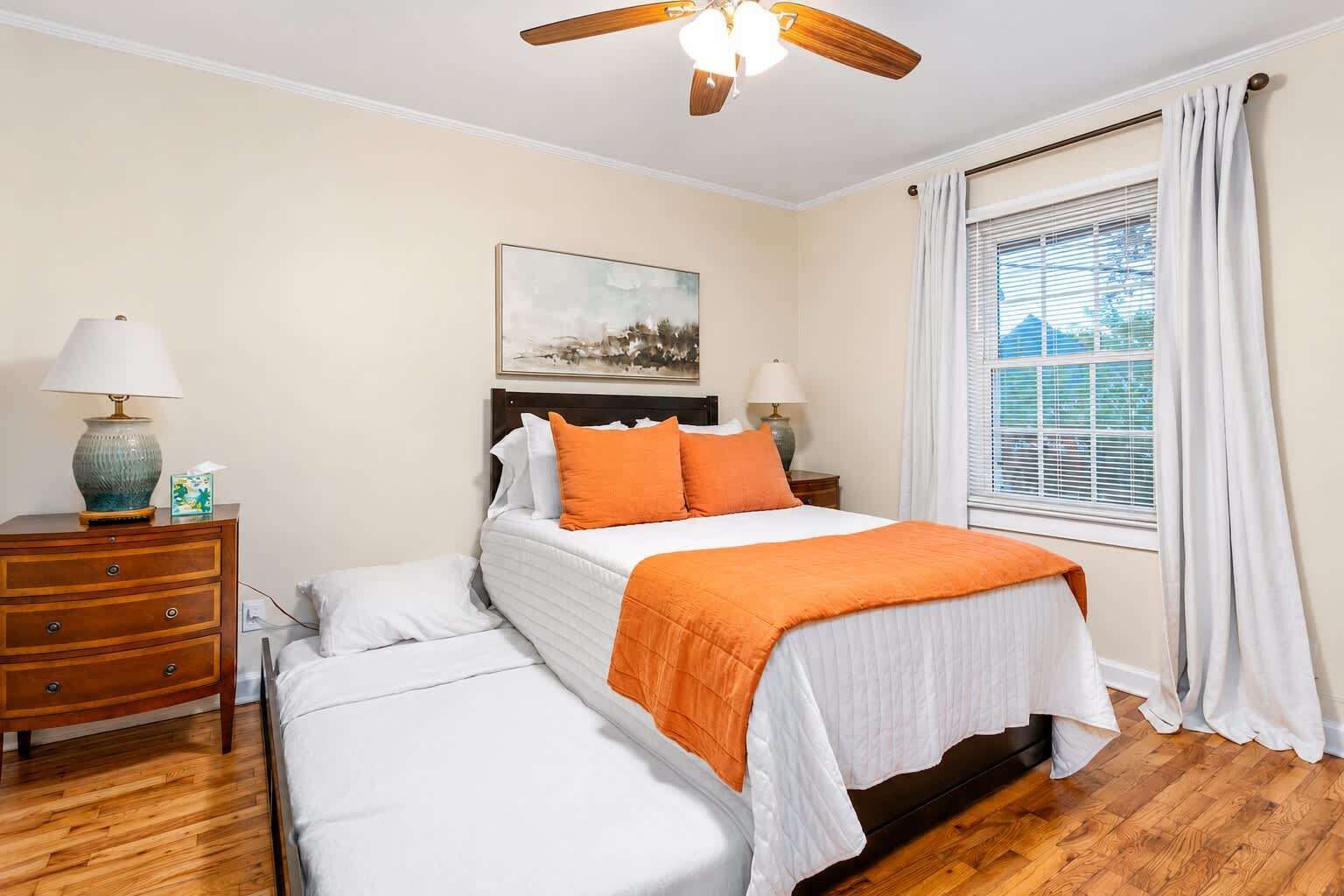 Une chambre à coucher comprenant un lit, un banc, une table de chevet, une lampe, un ventilateur de plafond, une fenêtre avec des rideaux et un tableau accroché au mur.