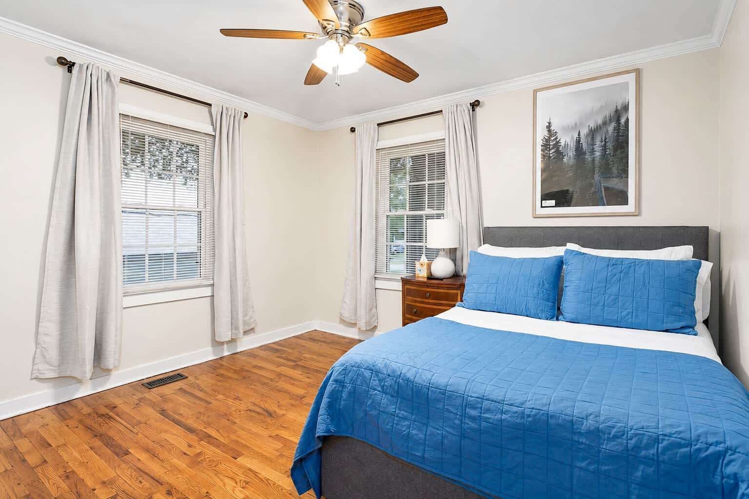 Une chambre à coucher avec un lit, un ventilateur de plafond, deux fenêtres avec des rideaux, une table de chevet avec une lampe et un tableau encadré accroché au mur.
