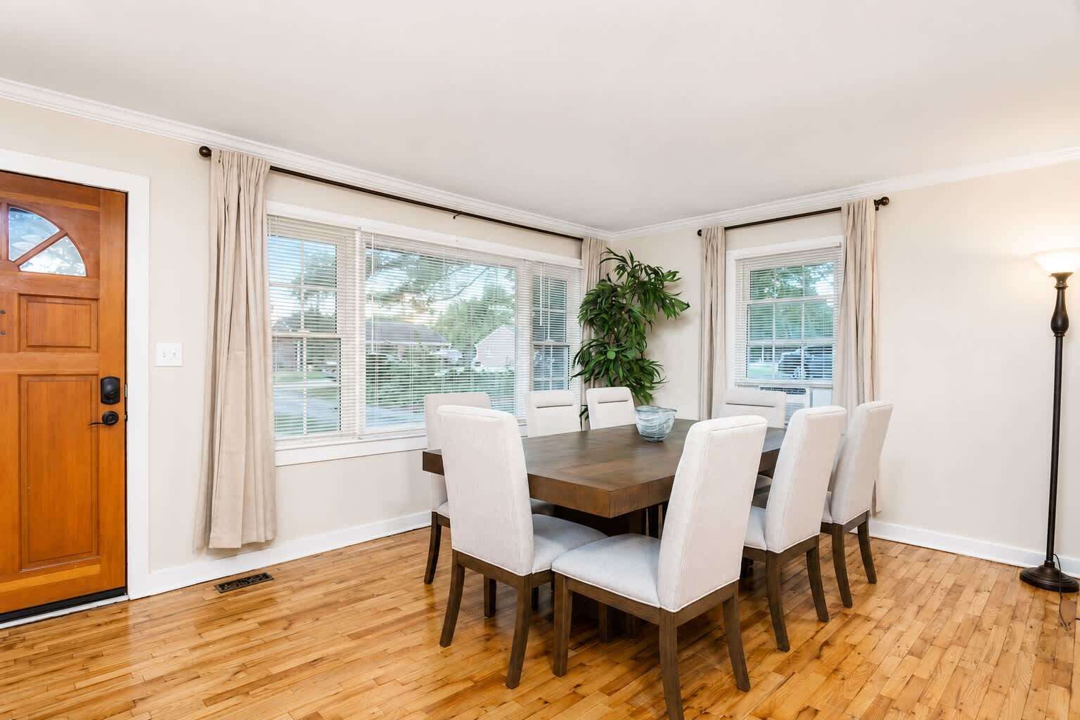 A dining room with a wooden table and white chairs, large windows with blinds, a potted plant, and a wooden door.