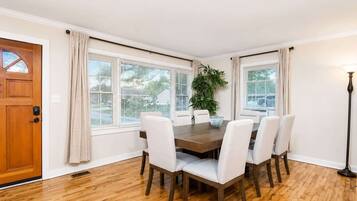 A dining room with a wooden table and white chairs, large windows with blinds, a potted plant, and a wooden door.