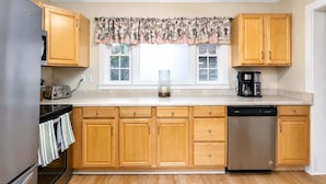 Una cocina con armarios de madera, lavavajillas de acero inoxidable, un tostador y un ventanal con un valance decorativo.