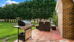 Un patio al aire libre con una parrilla y zona de estar, rodeado de árboles y un muro de ladrillo.