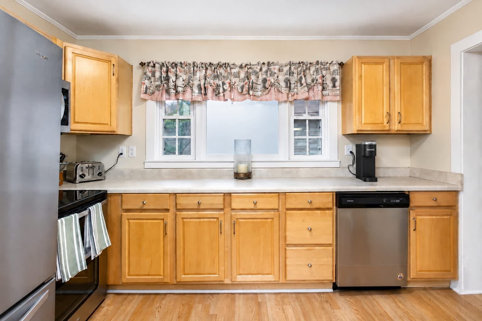 A kitchen with wooden cabinets, a stainless steel dishwasher, and a window with a patterned valance.