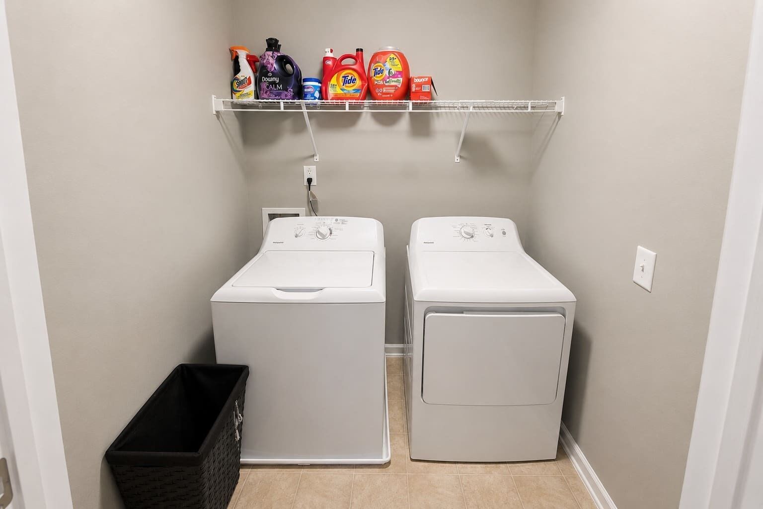 A laundry room with two white front-loading washing machines and a shelf above containing various cleaning products.