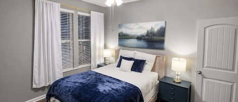 A bedroom with a bed, a nightstand, a ceiling fan, a window with blinds, and a door.