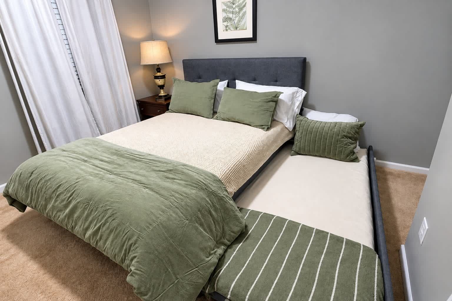 Een tweepersoonsbed met een grijs hoofdbord, twee kussens, een nachtkastje met een lamp en een ingelijst beeld aan de muur.