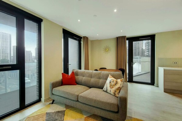 A modern living room with a sofa, a dining table, and large windows offering a city view. - Madison  (Wembley)