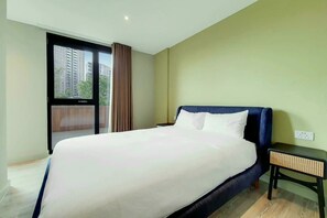 A hotel room with a large bed, a nightstand, a window with a view of buildings, and a ceiling light. - Madison  (Wembley)