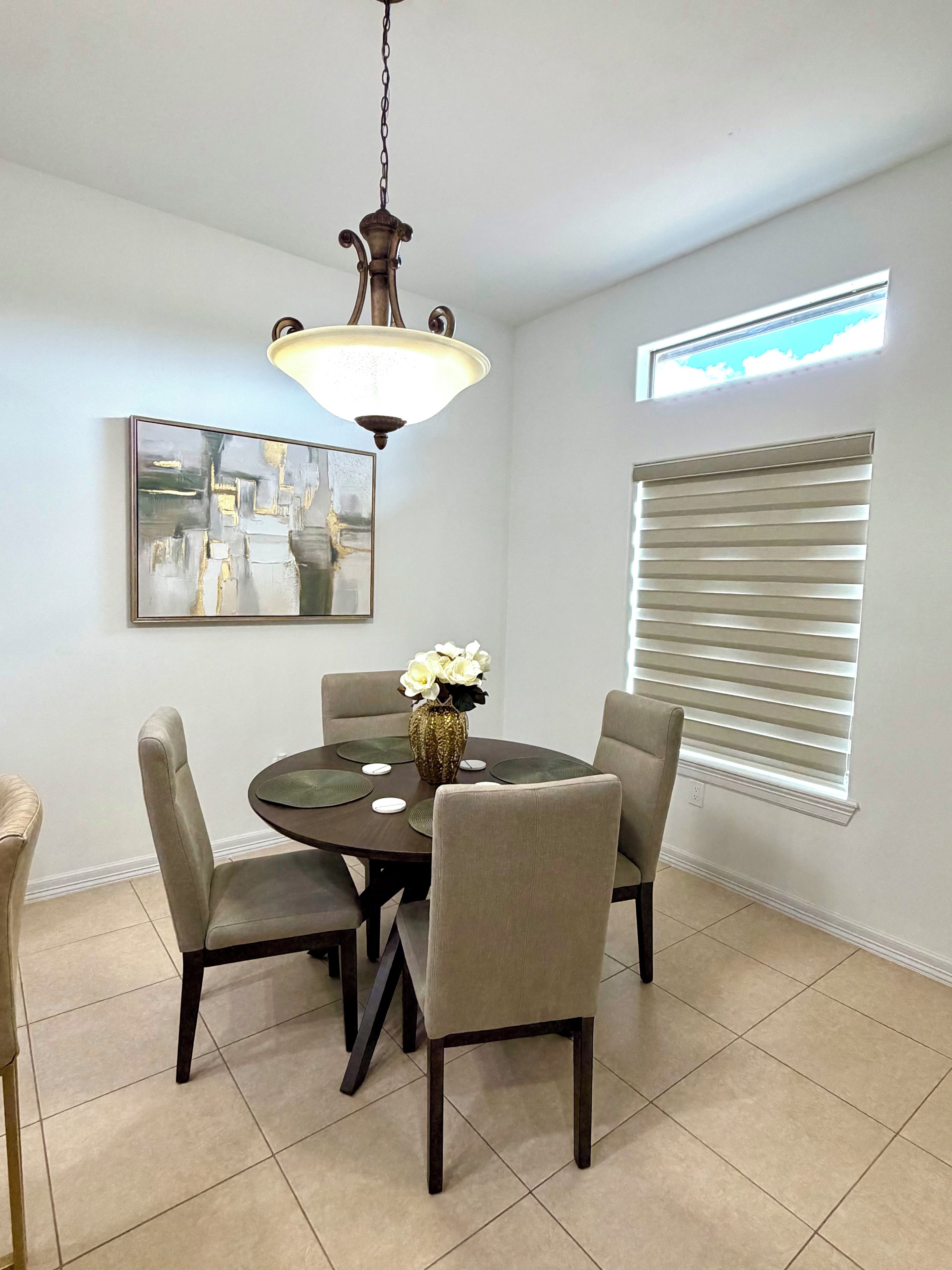 A dining area with a round table, four chairs, a vase with flowers, a ceiling light fixture, a wall-mounted painting, and a window with blinds.