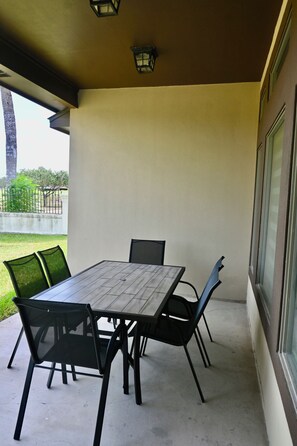 Ein überdachter Patio-Bereich mit Tisch und Stühlen, Blick ins Freie und ein Fenster auf der rechten Seite.