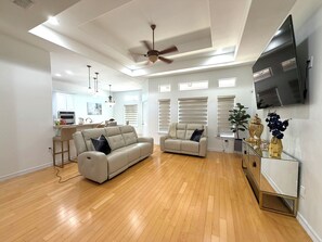 Ein geräumiger Wohnbereich mit Holzboden, zwei Sofas, einem Deckenventilator und einer Küche im Hintergrund.
