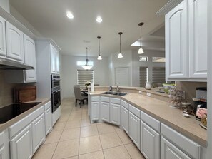 A modern kitchen with white cabinetry, a central island, pendant lighting, and a dining area with a table and chairs. - The Pinehurst Retreat - 3BR with Golf Course Views (Harlingen)
