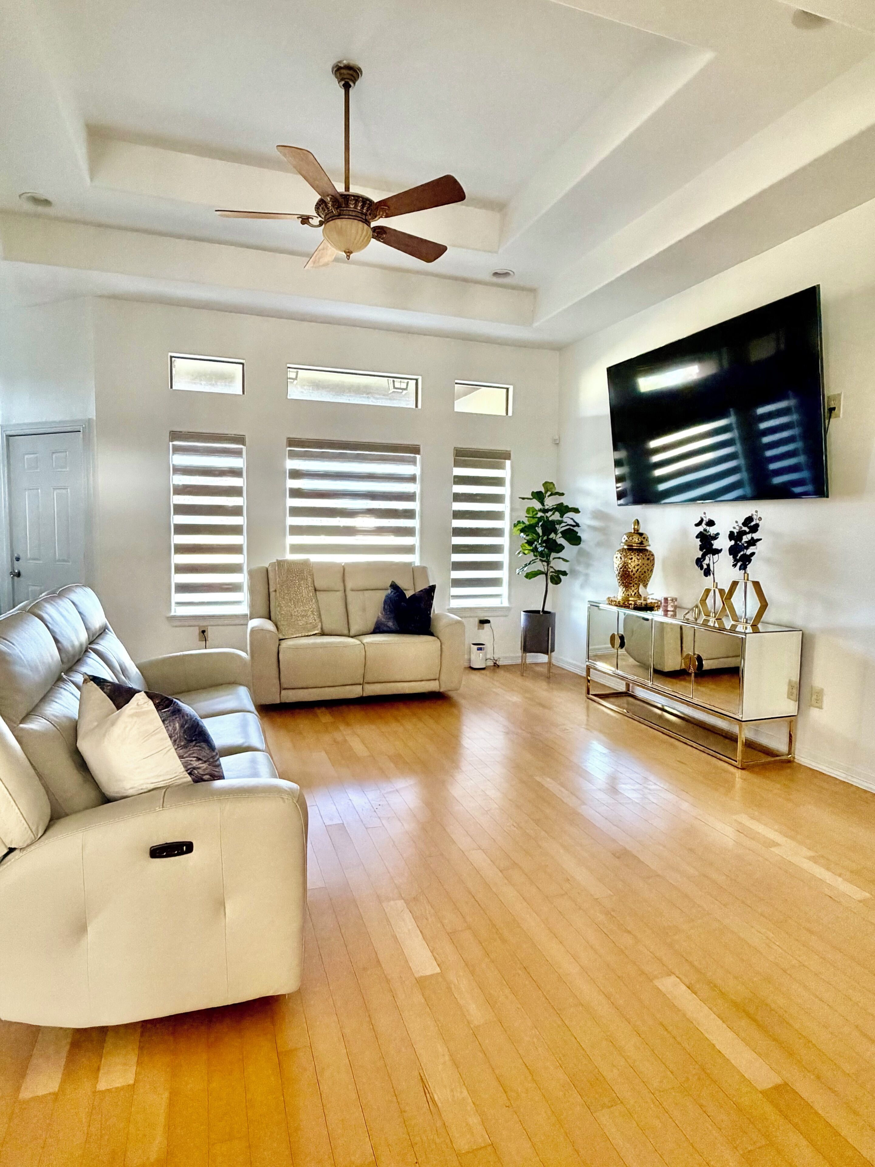 A spacious living room with a ceiling fan, a large flat-screen TV mounted on the wall, and a sofa with a decorative console table.