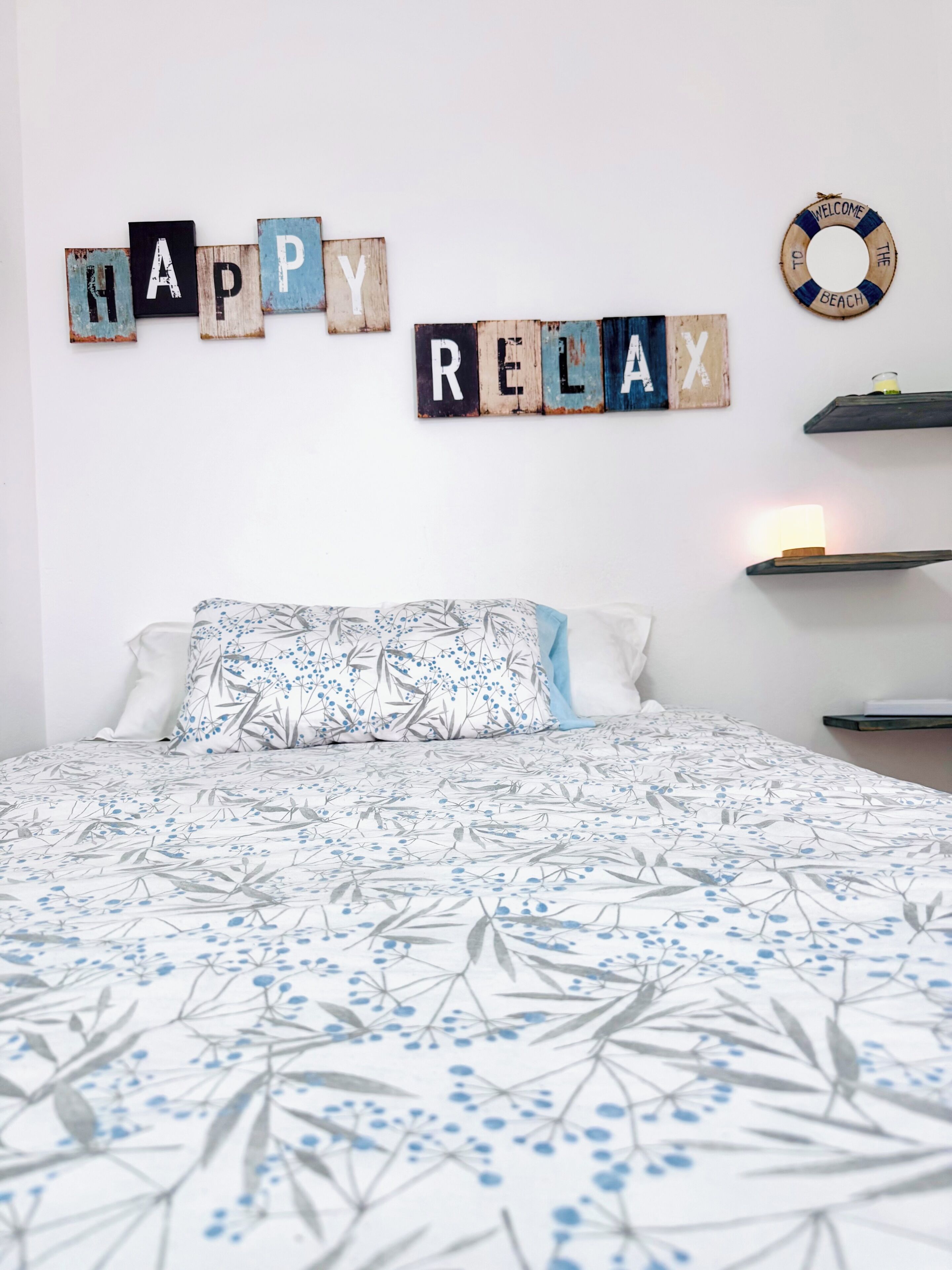 Un dormitorio con cama, almohadas, un cartel en la pared que dice 'HAPPY RELAX', un estante pequeño con una vela y un letrero que dice 'BIENVENIDOS A LA PLAYA'.